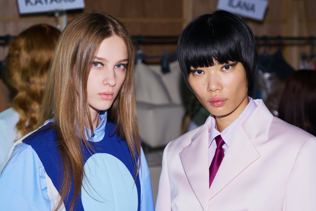 ファッションショーのバックステージで、2人の女性モデルがカメラを見つめています。一人は青いトップス、もう一人はピンクのスーツにタイを着用し、それぞれの個性が際立つスタイルです。