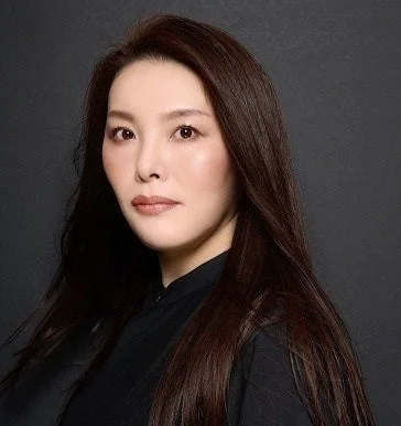 暗い背景の前で、黒いシャツを着た長い黒髪のアジア人女性のポートレート。彼女はカメラに向かって真剣な表情をしています。