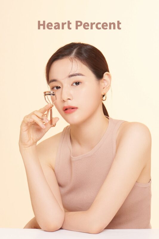 A model applying the eyelash curler to her lashes, with the Heart Percent logo