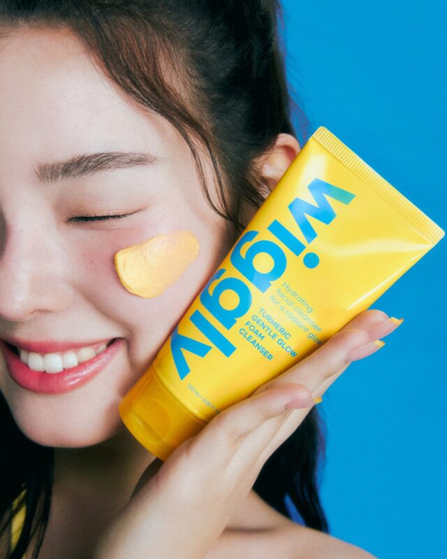 A woman holding a yellow tube with cream on her cheek; Turmeric Gentle Glow Foam Cleanser
