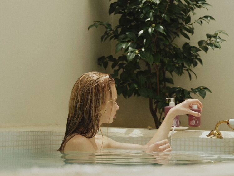 A woman with wet hair holding an ANILLO bottle in a bathtub